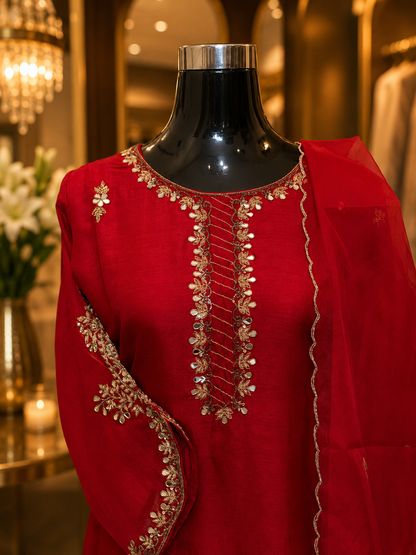 A bright red traditional outfit displayed on a black mannequin in an indoor boutique setting. The garment features ornate gold embroidery and mirror work around the neckline and along the draped dupatta.